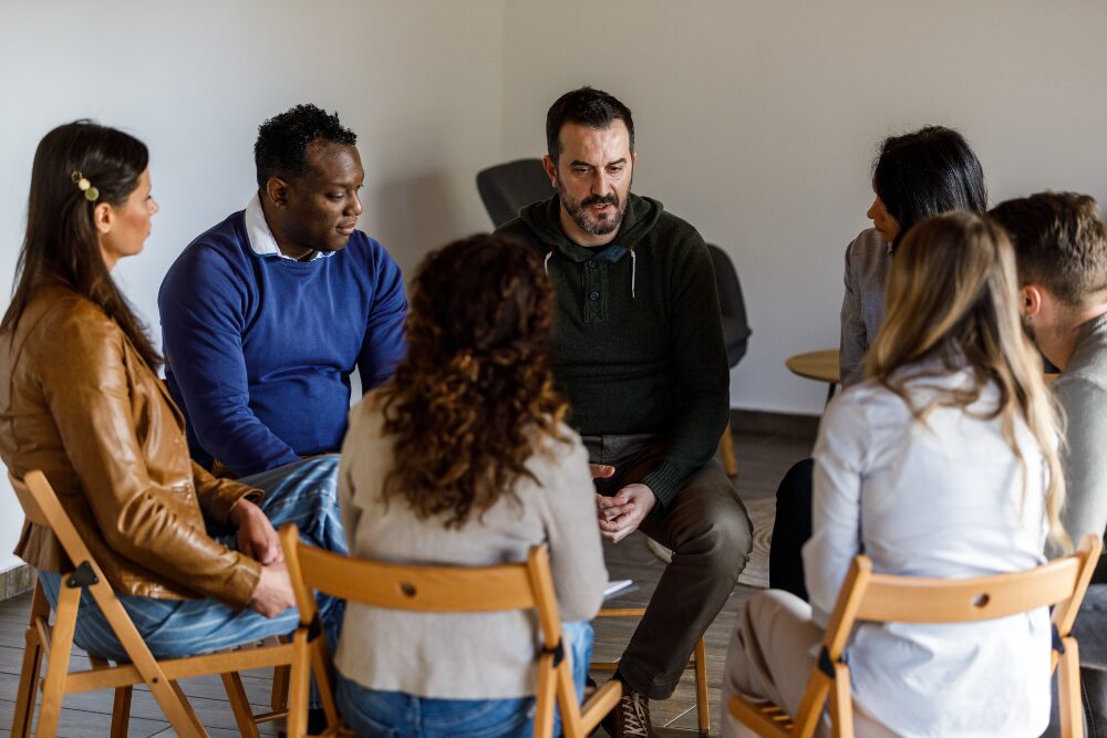 Group therapy for addiction treatment, showing adults in a supportive peer discussion led by a counselor.