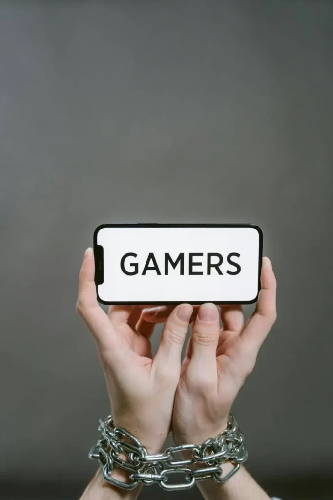 Illustration of hands chained holding a phone displaying “GAMERS,” symbolizing behavioral addiction to gaming.
